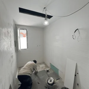 Un obrero arrodillado en un baño en construcción, colocando baldosas de color gris oscuro en el suelo. Las paredes están parcialmente alicatadas con azulejos blancos. Se ven herramientas, baldes y cables eléctricos colgando del techo.