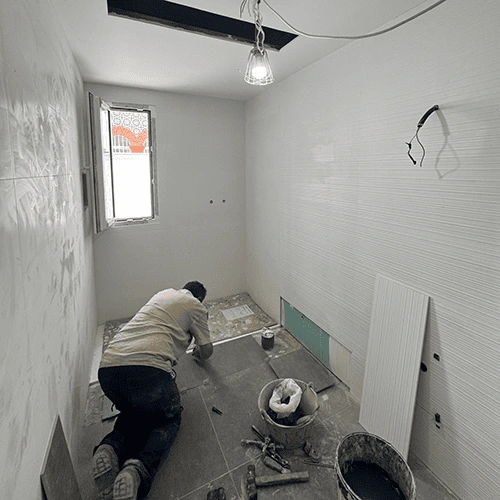 Fotografía de un baño en pleno proceso de reforma. Un trabajador, arrodillado, está colocando baldosas de color gris oscuro en el suelo. Las paredes están parcialmente alicatadas con azulejos blancos de textura horizontal. En el suelo, junto al obrero, hay herramientas y cubos de material de construcción. Del techo, con una abertura rectangular, cuelga una bombilla de obra.