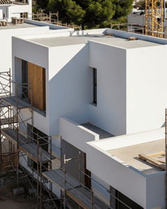Fondo con una imagen de una casa de nueva construcción en proceso, con andamios de metal rodeando la estructura. La casa, de estilo moderno y líneas cúbicas, está en gran parte enlucida de blanco. Una gran ventana con listones de madera verticales y otra ventana más pequeña y vertical se ven en la fachada principal. La construcción se encuentra en una zona con árboles y un andamio de grúa visible en el fondo. El cielo está despejado.