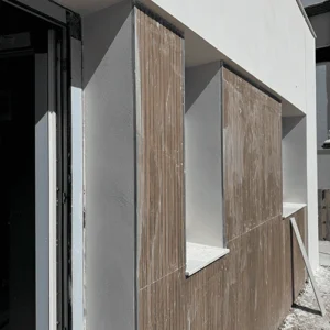 Primer plano de la fachada de una vivienda en fase de obra. La pared está revestida con paneles de madera de listones verticales y un acabado blanco en la parte superior. Hay dos huecos de ventana rectangulares y un marco de puerta de cristal en el lado izquierdo. La superficie de los paneles de madera está manchada por la obra y se aprecian restos de materiales en el suelo.