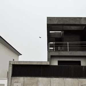 Vista del exterior de un edificio de diseño moderno en construcción. La estructura combina hormigón visto y grandes bloques de color negro. En el primer plano, hay una valla de hormigón con lamas metálicas verticales negras.