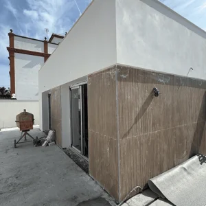 Fotografía del exterior de una vivienda en construcción con un diseño moderno. La fachada tiene un revestimiento de paneles de madera vertical en la parte inferior y paredes blancas en la superior. Delante de la casa, hay una solera de hormigón sin terminar. Se aprecian elementos de obra como una hormigonera oxidada, un rollo de material aislante y una puerta corredera de cristal.