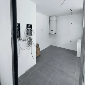 Una vista interior de una cocina-comedor moderna y luminosa. El espacio cuenta con una isla de cocina de color claro, una mesa de comedor de madera con un taburete alto gris. Al fondo, un gran ventanal ofrece vistas a un pequeño patio exterior con vegetación. La decoración es minimalista, con paredes blancas y suelo de color claro, y una lámpara de techo colgante sobre la mesa de comedor.