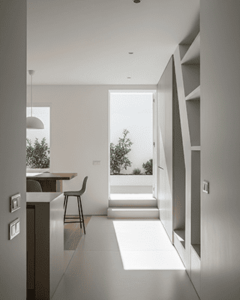 Fondo con una vista interior de un pasillo largo y minimalista con paredes blancas y un suelo claro. En la pared derecha, hay un mueble alto empotrado de madera con estanterías abiertas y espacios de almacenamiento. Al final del pasillo, se ve un patio exterior con paredes blancas y plantas verdes, al que se accede por unos escalones. La luz del sol ilumina el suelo, proyectando una sombra. A la izquierda, se puede ver parte de una cocina o comedor con una barra y un taburete.