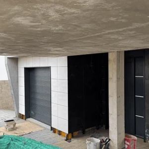Fotografía de un patio o zona exterior de una vivienda en fase de obra. Las paredes están revestidas con una combinación de azulejos grandes blancos y negros. A la izquierda se ve una persiana gris y a la derecha una puerta negra. El techo es de hormigón visto y se ven herramientas y materiales de construcción en el suelo.
