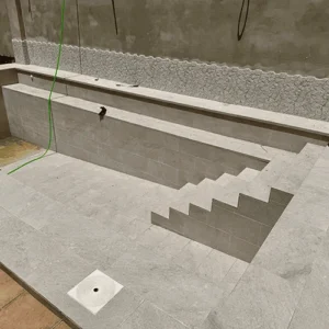 Fotografía de una piscina en proceso de construcción o reforma. El vaso de la piscina está vacío y tiene unas escaleras de obra revestidas con baldosas de piedra de color claro. El borde de la piscina también está alicatado con baldosas a juego. Se aprecian detalles de la obra como la estructura de hormigón, un cubo y una manguera en el interior.
