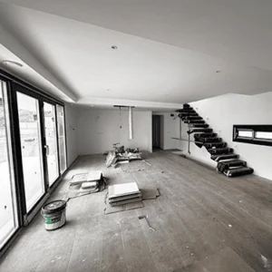Un salón diáfano en construcción con grandes ventanales de suelo a techo con marcos negros. El suelo es de madera inacabada. Al fondo, se ve una escalera de diseño moderno con peldaños negros. Hay cajas, azulejos y otros materiales de obra en el suelo.