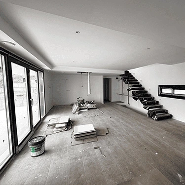 Un salón diáfano en construcción con grandes ventanales de suelo a techo con marcos negros. El suelo es de madera inacabada. Al fondo, se ve una escalera de diseño moderno con peldaños negros. Hay cajas, azulejos y otros materiales de obra en el suelo.