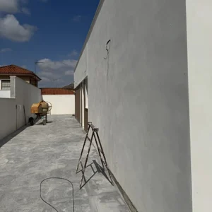 Una fotografía de una terraza o patio exterior en fase de construcción o reforma. El suelo está embaldosado con baldosas de color gris y las paredes son blancas. Una escalera de mano metálica se apoya en la pared de la derecha. En el fondo se distingue una hormigonera de cemento de color amarillo y los tejados de otras casas. El cielo es de color azul con algunas nubes.