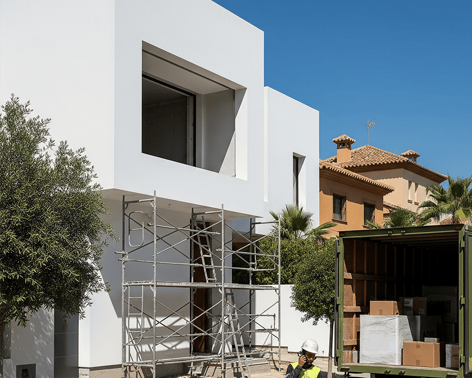 Fotografía del exterior de una vivienda moderna de color blanco en construcción, con andamios en la fachada. En primer plano, hay un hombre con casco de seguridad que habla por teléfono junto a la parte trasera de un camión de reparto. El camión tiene la puerta abierta, mostrando cajas en su interior.