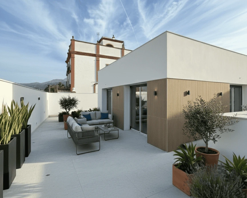 Terraza moderna y minimalista de un ático, con suelo de color claro, muebles de exterior contemporáneos y jardineras altas. El diseño combina paredes blancas lisas con revestimiento de madera clara. Al fondo, se aprecia la arquitectura tradicional andaluza.